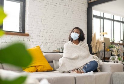 Upset multiracial woman with confirmed viral disease wearing medical mask feeling sad, sitting on the couch at home during quarantine and self isolation, taking measures to protect the family
